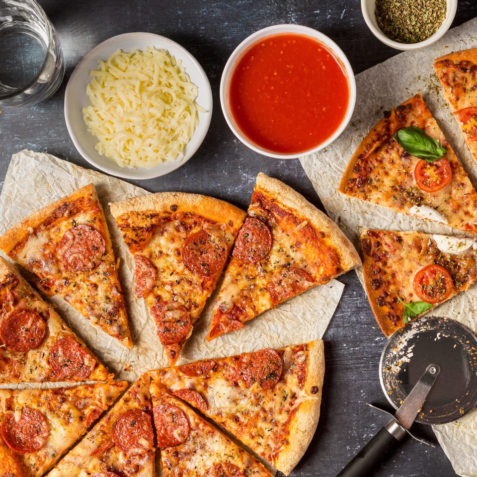 Overhead view of various pizza slices, cheese, sauce, and spices on a dark surface.