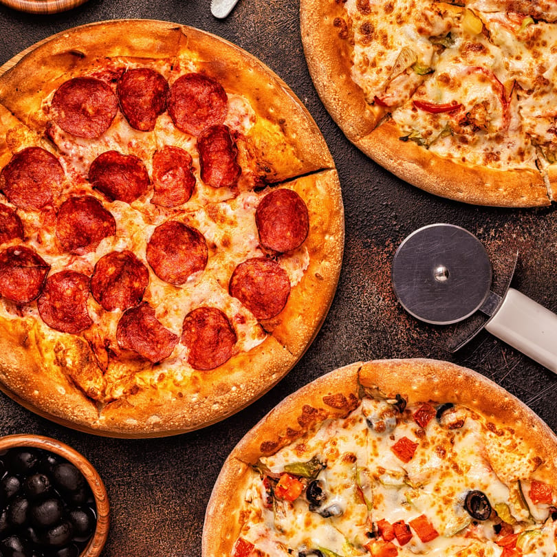 Three pizzas, including pepperoni and supreme, with a pizza cutter and bowl of olives on a dark surface.